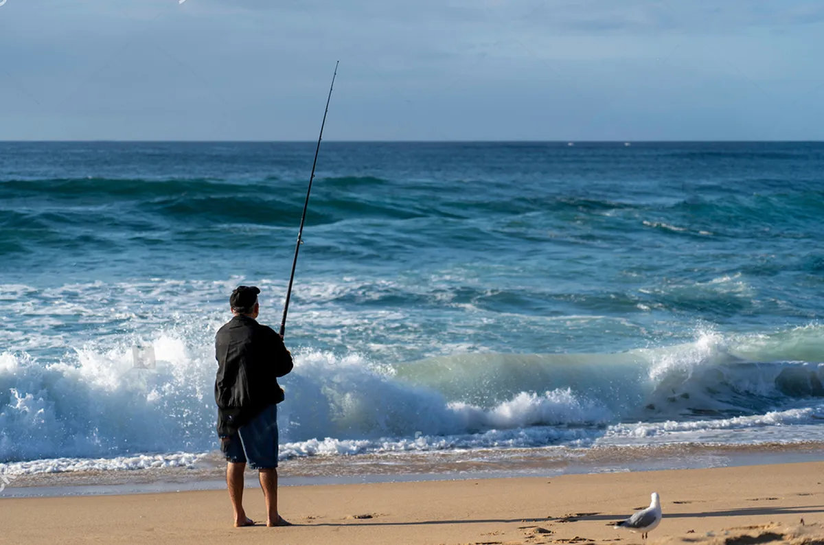 Ultimate Guide Beach Fishing: Tips and Tricks for Every Angler  2 A beach fisherman standing by the water, reeling in a line.